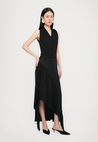 Black sleeveless dress with a V-neck, asymmetrical hemline, and draped fabric. Worn with pointed black heels and circular earrings.