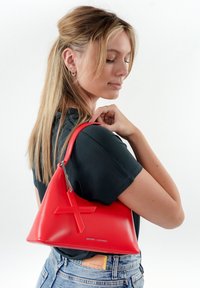 Bolso de mano triangular rojo con acabado mate, que presenta un destacado diseño cruzado y un detalle de cadena plateada. Llevado al hombro.