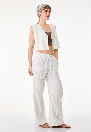 Junge Frau mit Kopftuch, die eine ärmellose, kurz geschnittene weiße Weste, ein braunes Crop-Top, lockere weiße Hosen mit Kordelzug und braune Sandalen trägt und vor einem weißen Hintergrund steht.