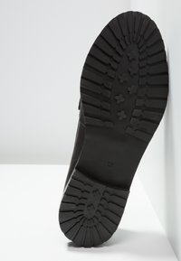 Semelle de botte noire avec des motifs de semelle profonds et taille 37 marquée, positionnée verticalement contre une surface et un mur blancs.