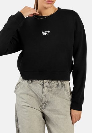 Vrouw draagt een zwart, cropped Reebok-sweatshirt en lichtgrijze, hooggetailleerde jeans, terwijl ze een gouden ketting dicht bij haar hals vasthoudt.