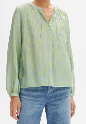 Femme portant un chemisier vert clair à manches longues avec des motifs jaunes subtils et un jean bleu, debout devant un fond uni.
