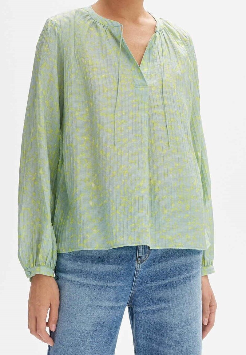Femme portant un chemisier vert clair à manches longues avec des motifs jaunes subtils et un jean bleu, debout devant un fond uni.