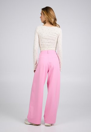 Vrouw met de rug naar de camera, gekleed in een crème kleurige top met lange mouwen en een kleine bloemenprint, gecombineerd met hoog getailleerde, brede roze broek en witte sneakers.