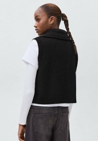 Frau mit geflochtenem Haar trägt einen schwarzen ärmellosen Pullover über einem weißen Langarmshirt und dunklen Jeans, sie schaut nach links vor einem neutralen Hintergrund.