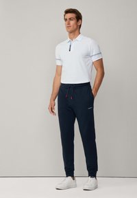 Weißes Polohemd mit Reißverschlusskragen und marineblauen Bündchen, kombiniert mit marineblauen Jogginghosen mit Kordelzug und Logo. Dazu weiße Sneaker.