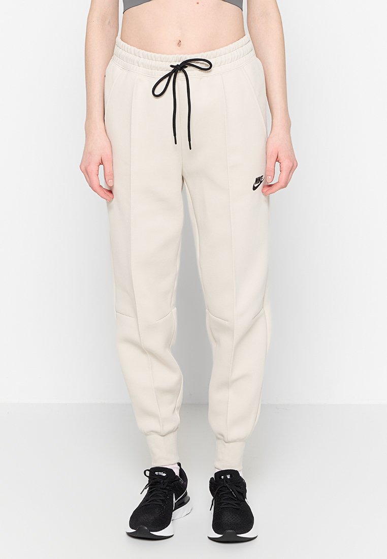 Pantalones deportivos beige con cintura elástica, cordón negro, bolsillos laterales y un pequeño logo negro de Nike en la parte superior del muslo.