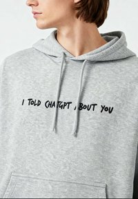 Grijze hoodie met een voorste kangoeroezak, capuchon met trekkoord en geborduurde tekst in het zwart met de tekst "IK HEB CHATGPT OVER JOU GEPRAAT."
