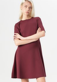 Rochie burgundy cu mânecă scurtă, fabricată dintr-un material moale, cu decolteu rotund, fustă evazată și design minimalist. Textură netedă cu o siluetă curată.