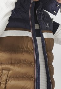 Brown and navy puffer vest featuring horizontal white stripes, zip closure, and high collar. Textured fabric with a quilted design.