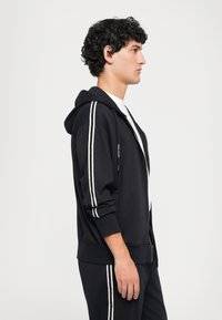Black zip-up hoodie with white side stripes, adjustable drawstring hood, and ribbed cuffs, paired with matching pants.