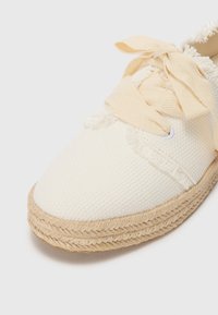 Hvit lerret espadrille med flisede kanter, beige snører og en vevd jutesåle. Teksturen virker myk med en strukturert form.