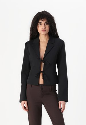 Black cropped blazer with long sleeves, front tie closure, and notched collar, paired with fitted brown pants featuring two buttons.