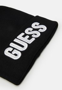 Berretto di lana nera con un logo bianco in rilievo "GUESS" decorato con borchie metalliche, caratterizzato da una finitura aderente e testurizzata.
