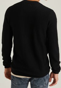 Maglione nero a maglia con maniche lunghe, caratterizzato da una superficie texturizzata e un collo a giro, indossato sopra una camicia di colore chiaro e jeans in denim.
