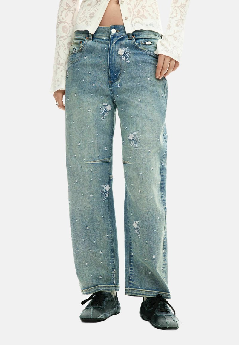 Naine, kes kannab helehalli, kulunud laia säärega teksaseid väikeste valgete tikitud lillidega ja musti paeltega kingi.