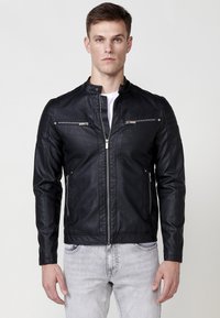 Chaqueta de cuero negra con cuello alto, cremallera frontal y dos bolsillos en el pecho con cremallera. Presenta detalles acanalados en los hombros. Textura suave.
