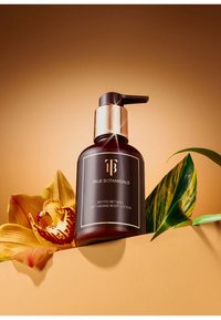 Flacon de lotion en verre brun foncé avec une pompe dorée, étiqueté "True Botanicals Phyto-Retinol Lotion Corporelle Anti-Âge", sur un fond brun et une fleur.