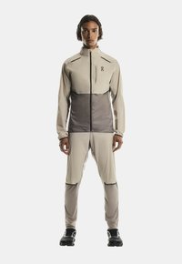 Personne debout de face portant une veste et un pantalon de sport beiges et gris avec des chaussures de course noires sur fond blanc.