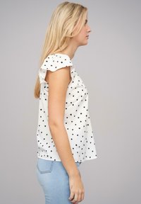 Weiße Bluse mit schwarzem Herzmuster, kurzen Ärmeln und lockerer Passform. Kombiniert mit hellblauen Jeans; glattes Material, schlichtes Design.