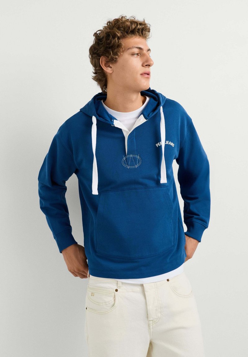 Junger Mann mit lockigem Haar, der einen blauen Hoodie mit weißen Kordeln und cremefarbenen Hosen trägt und seitlich vor einem einfarbigen Hintergrund schaut.