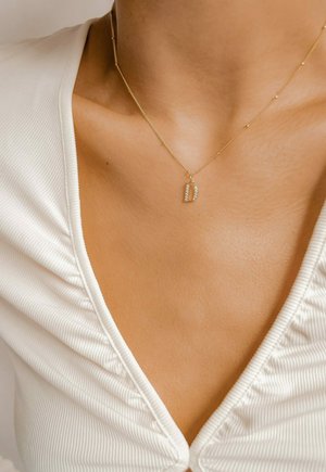 Gouden ketting met een kleine, schitterende letter 'D' hanger versierd met heldere stenen, gelegen op een gestructureerde witte stoffen top.