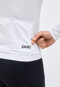 Χέρι που τραβάει μικρό φερμουάρ στην πίσω τσέπη ενός λευκού μακρυμάνικου ποδηλατικού τζέρσεϊ με το λογότυπο "poc", φορώντας μαύρο παντελόνι.