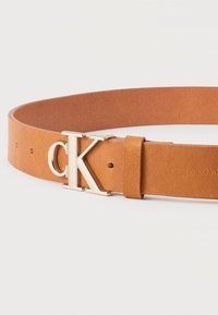 Ταμπά δερμάτινη ζώνη με ασημί αγκράφα "CK" και δερμάτινη θηλιά, παρουσιασμένη πάνω σε λευκό απλό φόντο.