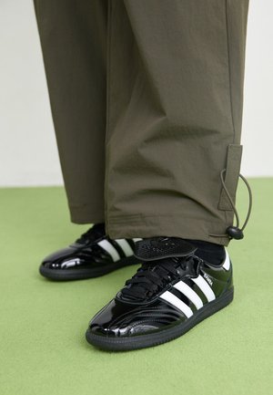 Sneakers in vernice nera con tre strisce bianche, lacci neri e suola nera testurizzata, abbinati a pantaloni verde oliva.