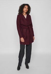 Burgundijas apvalks ar plašu apkakli, jostu viduklī un garām piedurknēm, apvienots ar melniem biksēm un potīšu zābakiem. Gluda tekstūra un pielāgota piegriezība.