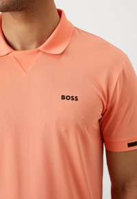 Rosa polo skjorte laget av teksturert stoff, med en svart "BOSS"-logo på venstre bryst og en svart stripe på ermet.