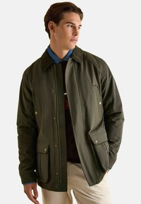 Zielona oliwkowa kurtka z zamkiem z przodu, dwoma kieszeniami z klapkami i teksturowanym kołnierzem. Posiada gładkie wykończenie i kontrastującą niebieską warstwę wewnętrzną.