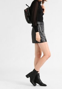 Minirock aus schwarzem Leder mit silbernen Ösen, kombiniert mit einem schwarzen Langarmshirt und Stiefeletten. Dazu ein dekorativer schwarzer Rucksack.