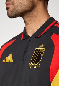 Camisa polo preta com detalhes em vermelho e amarelo, apresentando um bolso com fecho, logótipo bordado e três riscas amarelas no ombro.