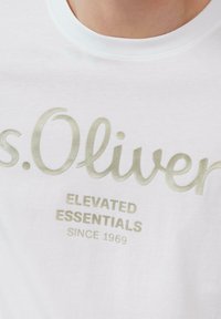 Weißes Baumwoll-T-Shirt mit kurzen Ärmeln. Mit metallic-goldenem Text "s.Oliver" und "ELEVATED ESSENTIALS SINCE 1969" darunter bedruckt. Glatte Textur.