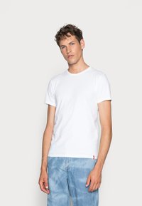 Hvid bomulds t-shirt med korte ærmer, rund halsudskæring og et vævet labeldetalje ved kanten. Båret med lyse denimjeans.