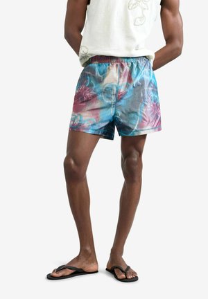 Uomo che indossa pantaloncini da bagno floreali multicolori blu e rosa, canottiera bianca e infradito nere, in piedi con le mani dietro la schiena.