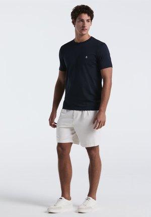 T-shirt bleu marine à manches courtes en tissu lisse, associé à un short blanc et des baskets blanches, avec un petit logo sur la poitrine.