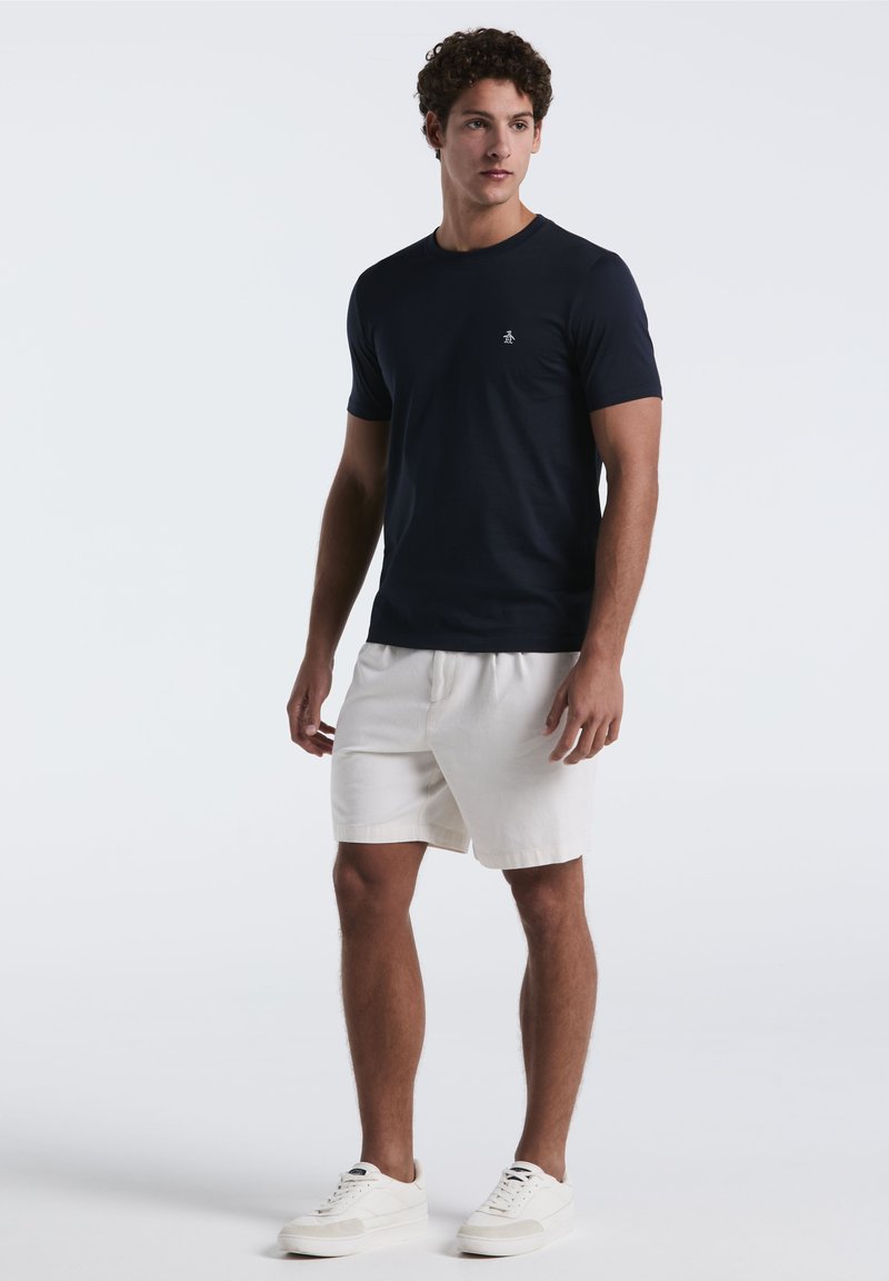 T-shirt bleu marine à manches courtes en tissu lisse, associé à un short blanc et des baskets blanches, avec un petit logo sur la poitrine.