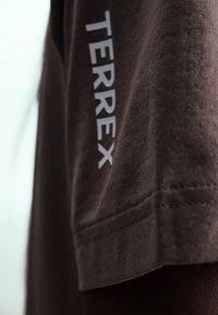 Gros plan sur un tissu marron avec le texte blanc « TERREX » imprimé verticalement près de l'ourlet et détails de couture visibles.