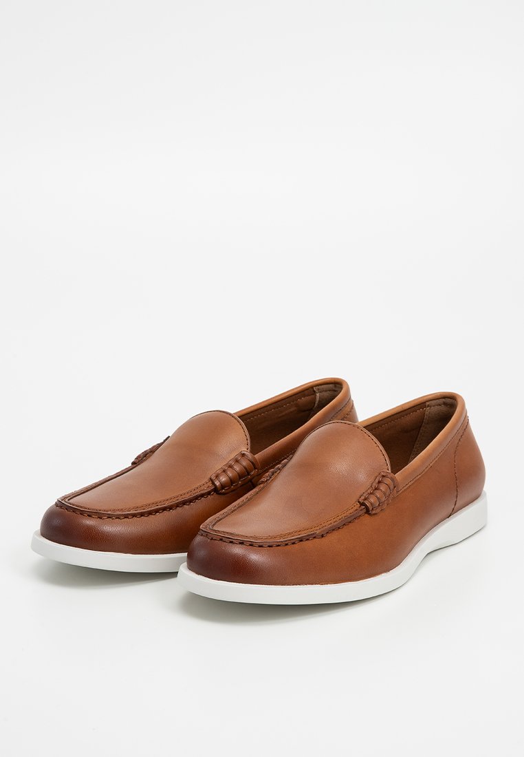 Des chaussures en cuir marron à enfiler présentent un bout rond, des coutures intriquées et une semelle en caoutchouc blanc pour le contraste. Texture lisse sans motifs visibles.