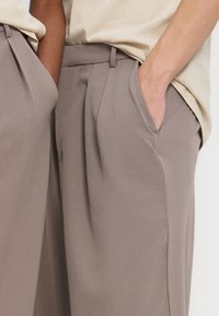 Δύο άτομα στέκονται δίπλα δίπλα φορώντας μπεζ τοπ και ταουπ παντελόνια με ψηλή μέση, με τα χέρια στις πλαϊνές τσέπες.