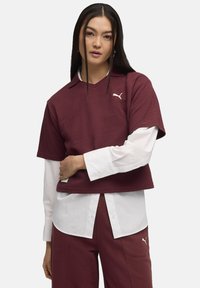 Maglietta polo a coste color bordeaux con maniche corte, indossata sopra una camicia bianca a maniche lunghe, caratterizzata da un logo sottile e una vestibilità rilassata.
