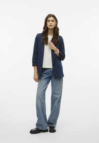 Blazer marin sur un t-shirt blanc, associé à un jean large bleu clair. La tenue comprend un bracelet et des chaussures noires avec un design texturé.