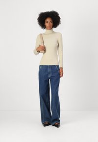 Lys beige turtleneck genser, løse blå denimjeans og svarte leiligheter. Modellen holder en beige skulderreimsekk mot en nøytral bakgrunn.