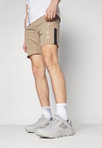 Beiga shorts med en marinblå sidopanel, dekorerande vita grafik och i ett lätt material, kombinerade med grå sportskor.