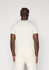 Cremfarbendes T-Shirt mit kurzen Ärmeln und rundem Halsausschnitt, mit einem kleinen schwarzen Akzent auf der Rückseite. Glatte Textur, enganliegendes Design, kombiniert mit passenden Hosen.