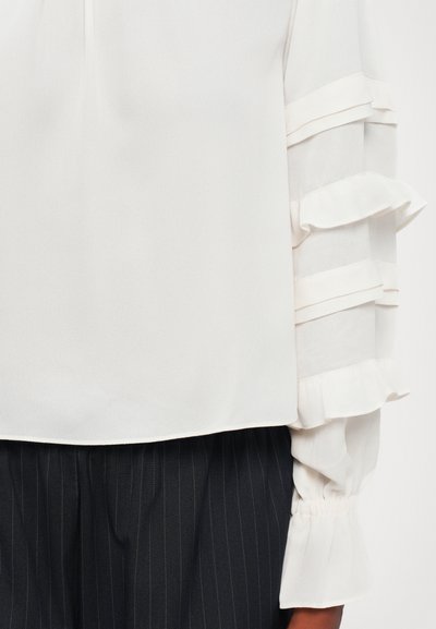 Blouse blanche avec des manches en tulle à volants, associée à un pantalon noir à rayures, présentée de la taille à mi-cuisse sur une personne.