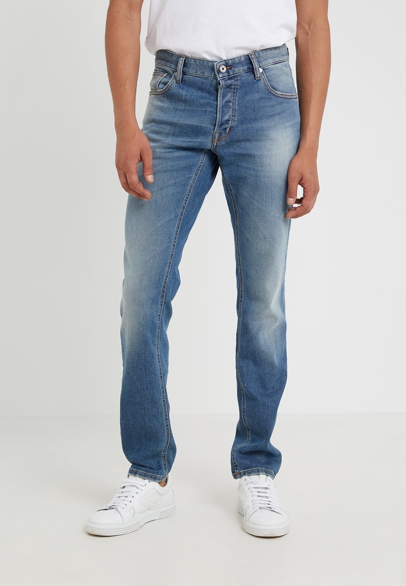Homme portant un jean bleu délavé avec des baskets blanches et une chemise blanche, debout devant un fond clair uni.