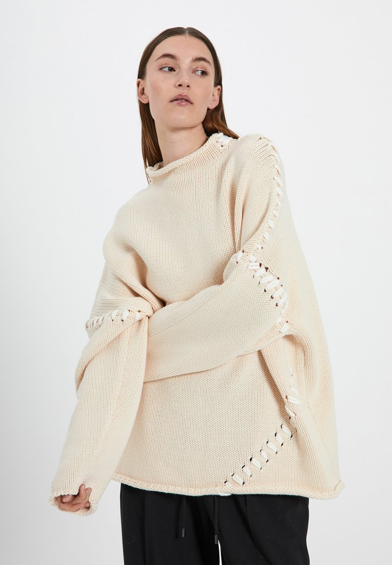 Suéter oversized de color crema con textura de punto abierto, detalle de encaje lateral y cuello alto, llevado sobre pantalones negros.